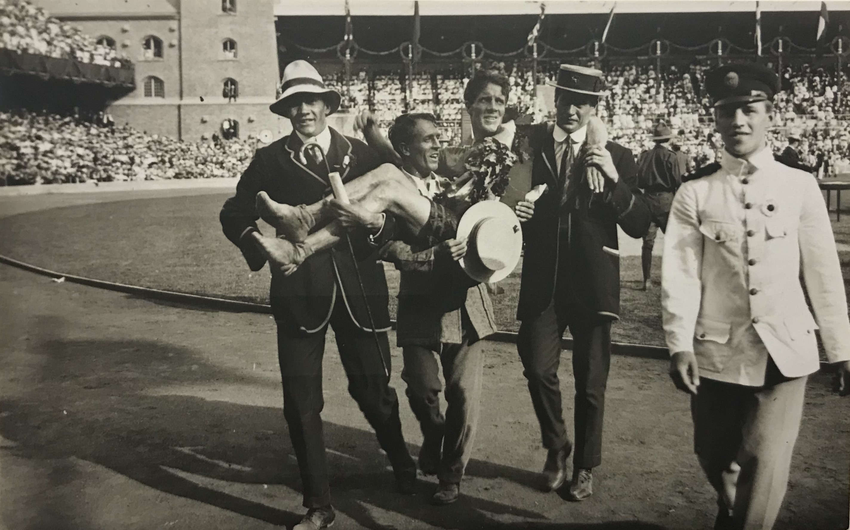 Svartvitt fotografi av en man med bara fötter och segerkrans som bärs av tre kostymklädda män inför fullsatta läktare på Stockhkolms stadion.