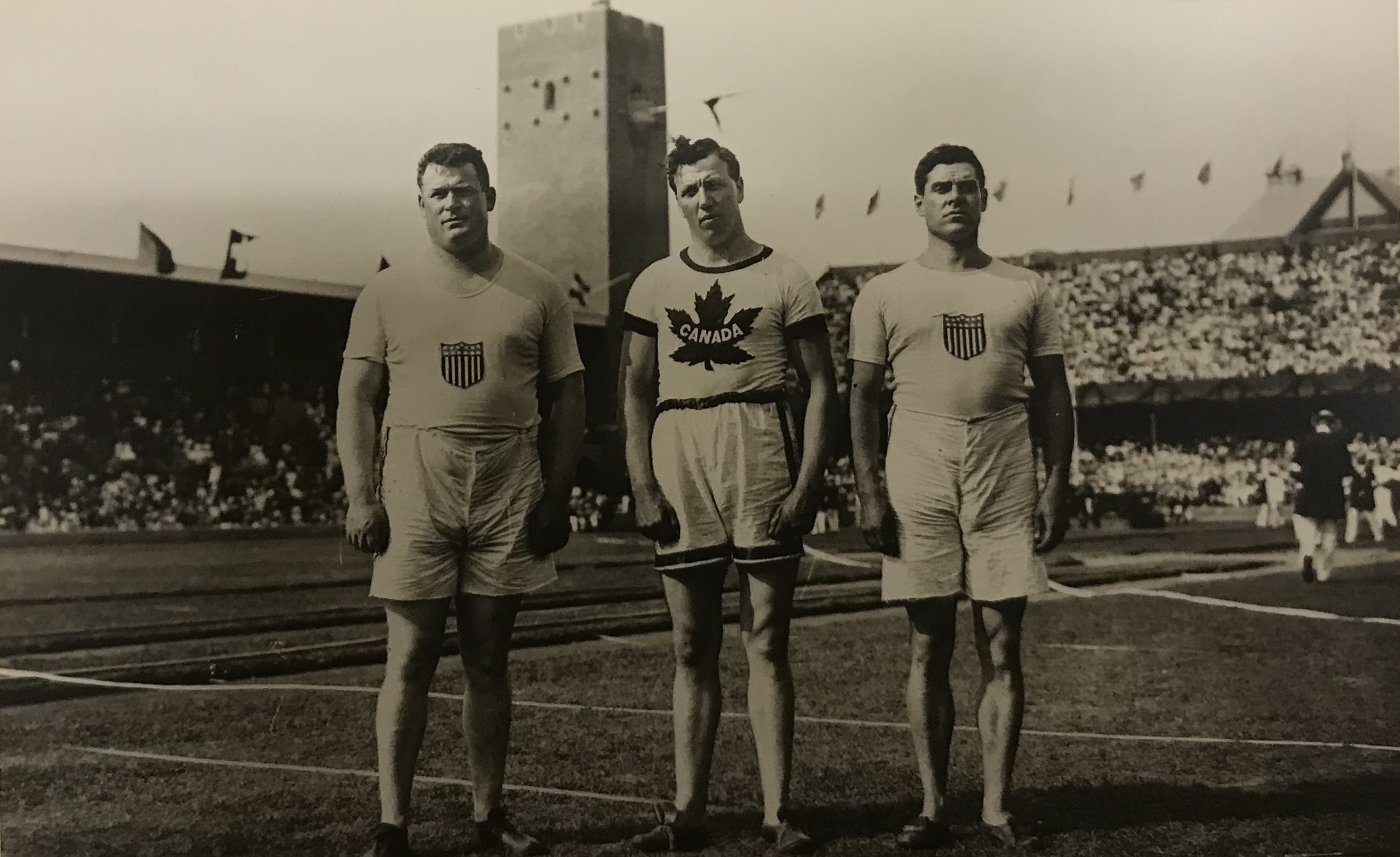 Svavrtvitt fotografi tre unga män klädda i shorts och t-shirts med amerikanska och kanadensiska emblem stående på gräsmattan på Stockholms stadion.