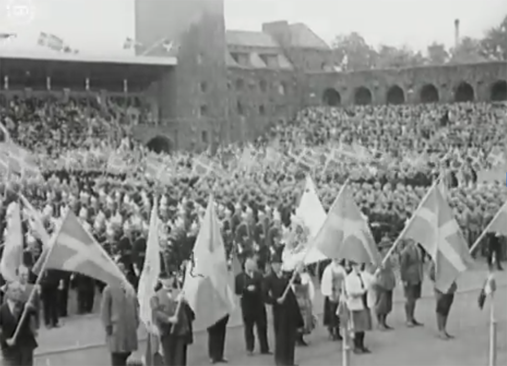 Svartvit bild av folksamling och flaggbärare på innergården av en stor byggnad. 