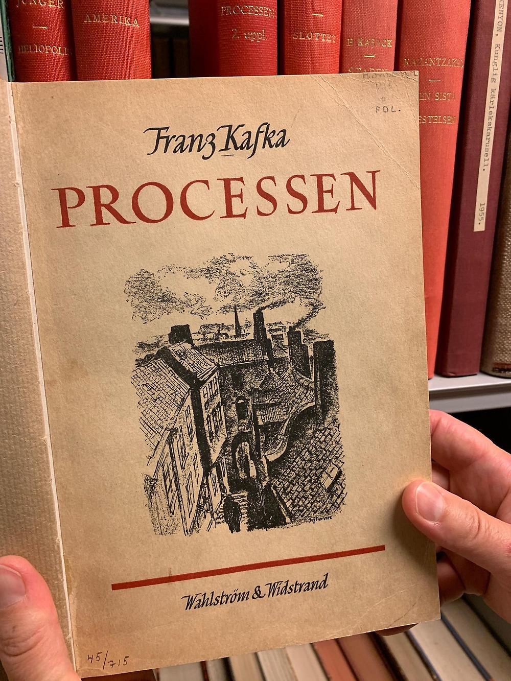 Händer håller i uppslagen bok framför hylla med röda bokryggar. På den uppslagna boken står det Processen i röd skrift och därunder finns en teckning i svart-vitt med hus och fabriker samt en gestalt i förgrunden.